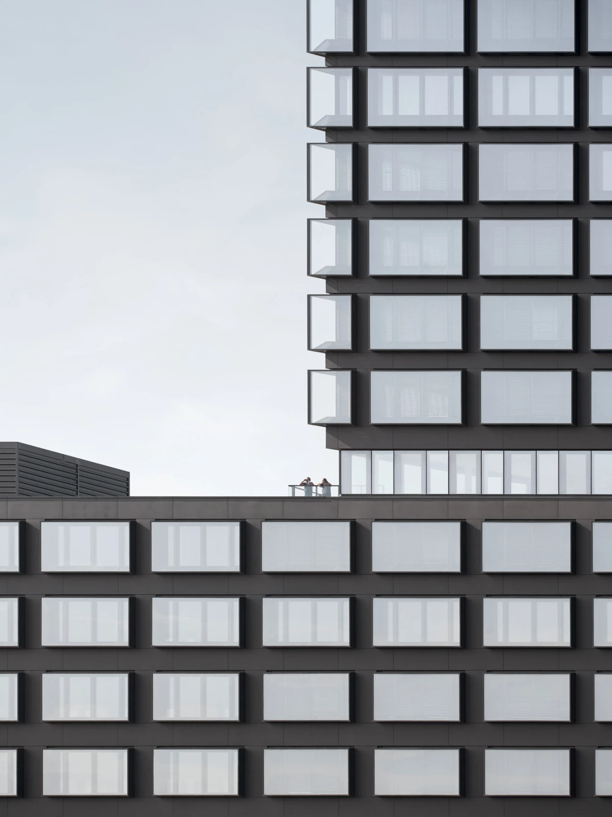 Two people stand on a rooftop terrace of a tall, modern glass-office building with a grid of windows.