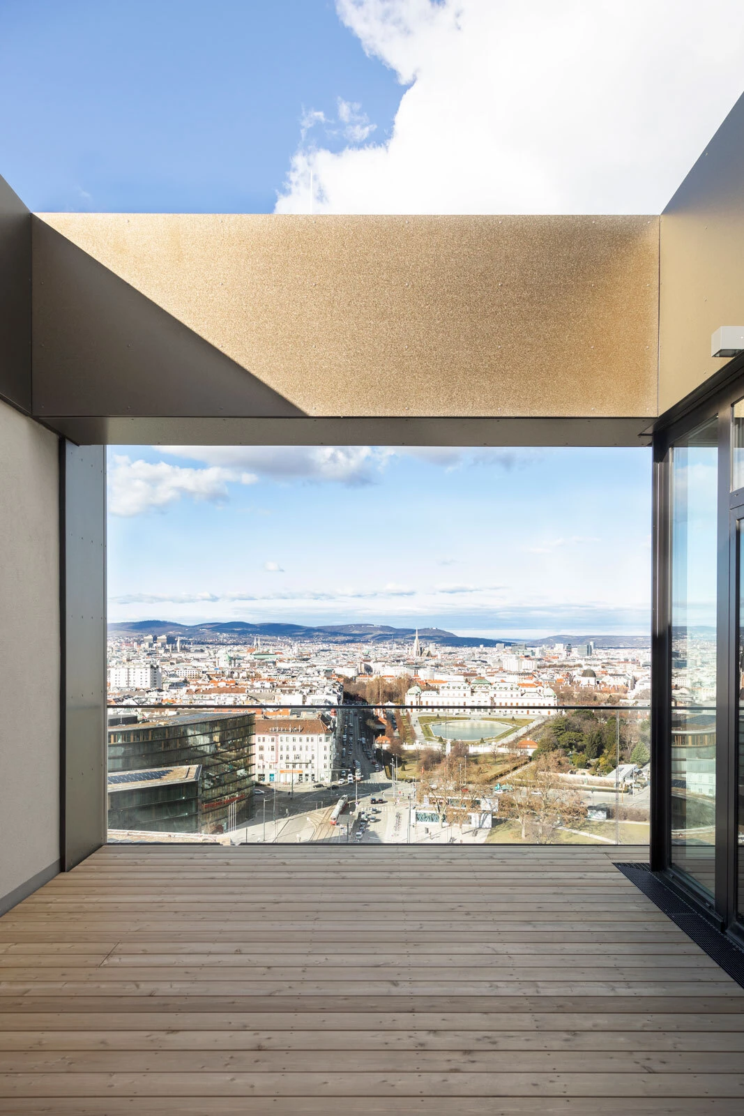 Wooden-deck balcony with glass railing overlooks a cityscape under a blue sky.