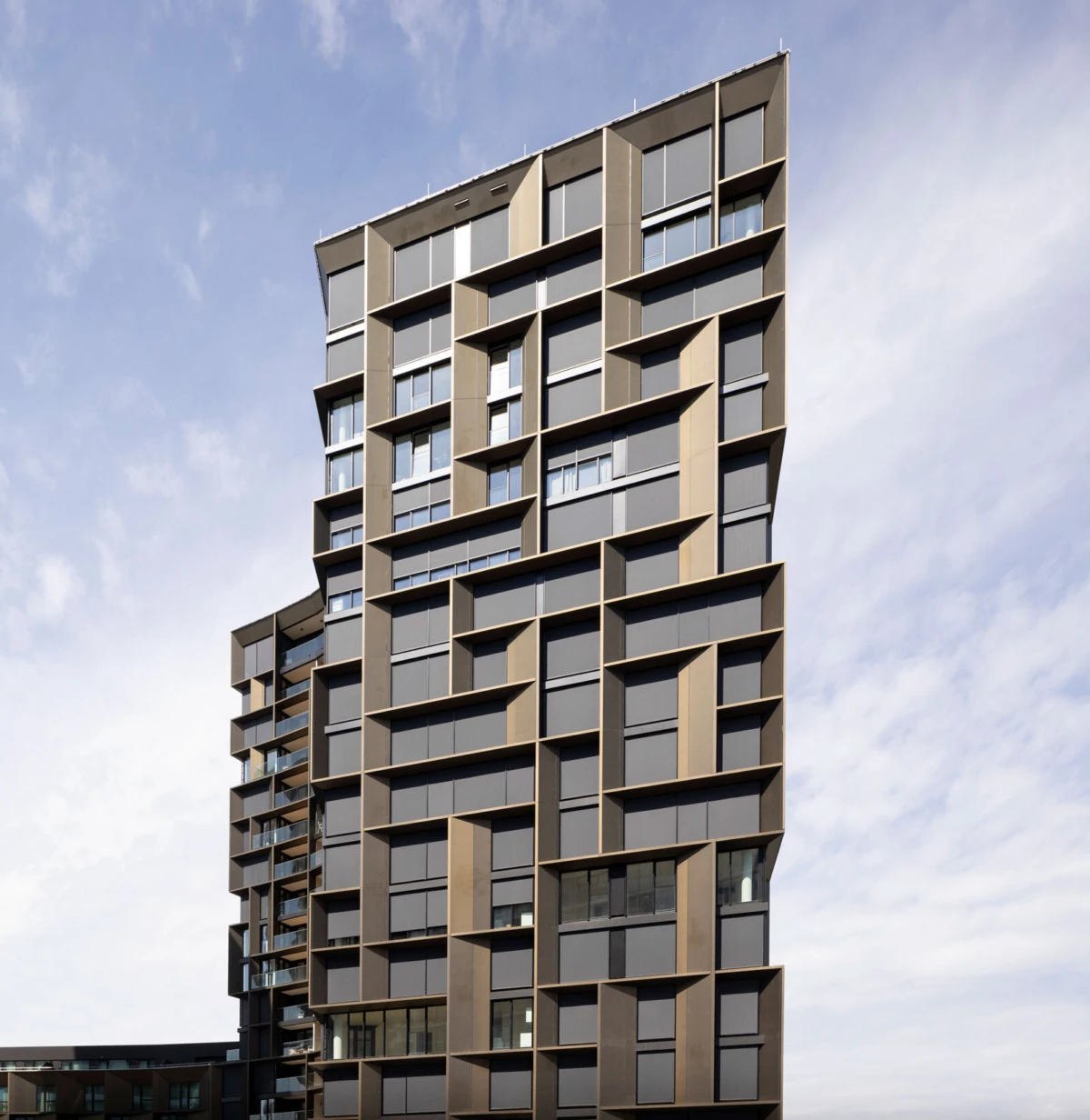 Tall modern high-rise apartment building with angular brown facade and grid of windows against a blue sky.