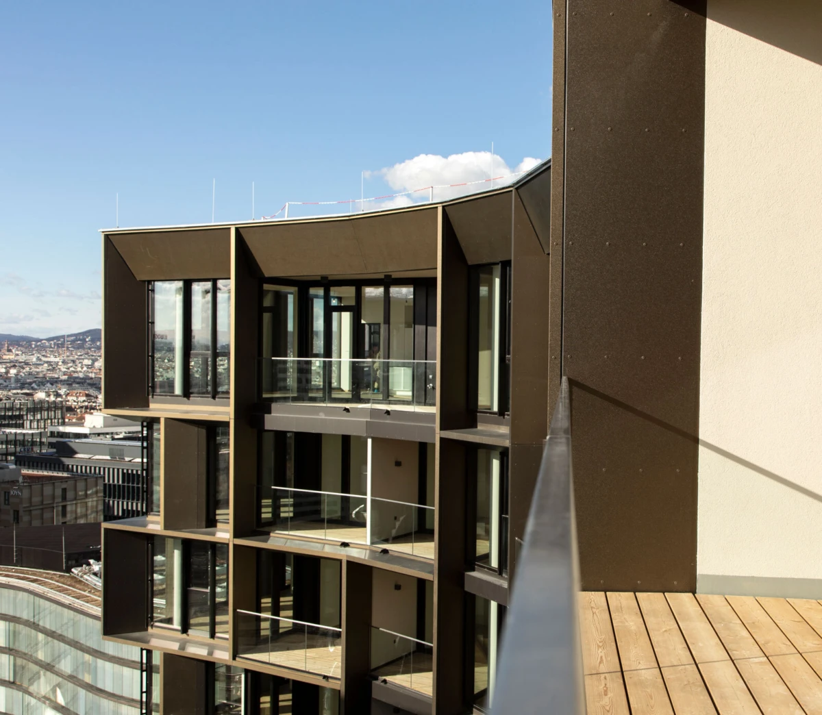 Brown glass-front apartment building with balconies, viewed from a wooden rooftop deck overlooking a cityscape.