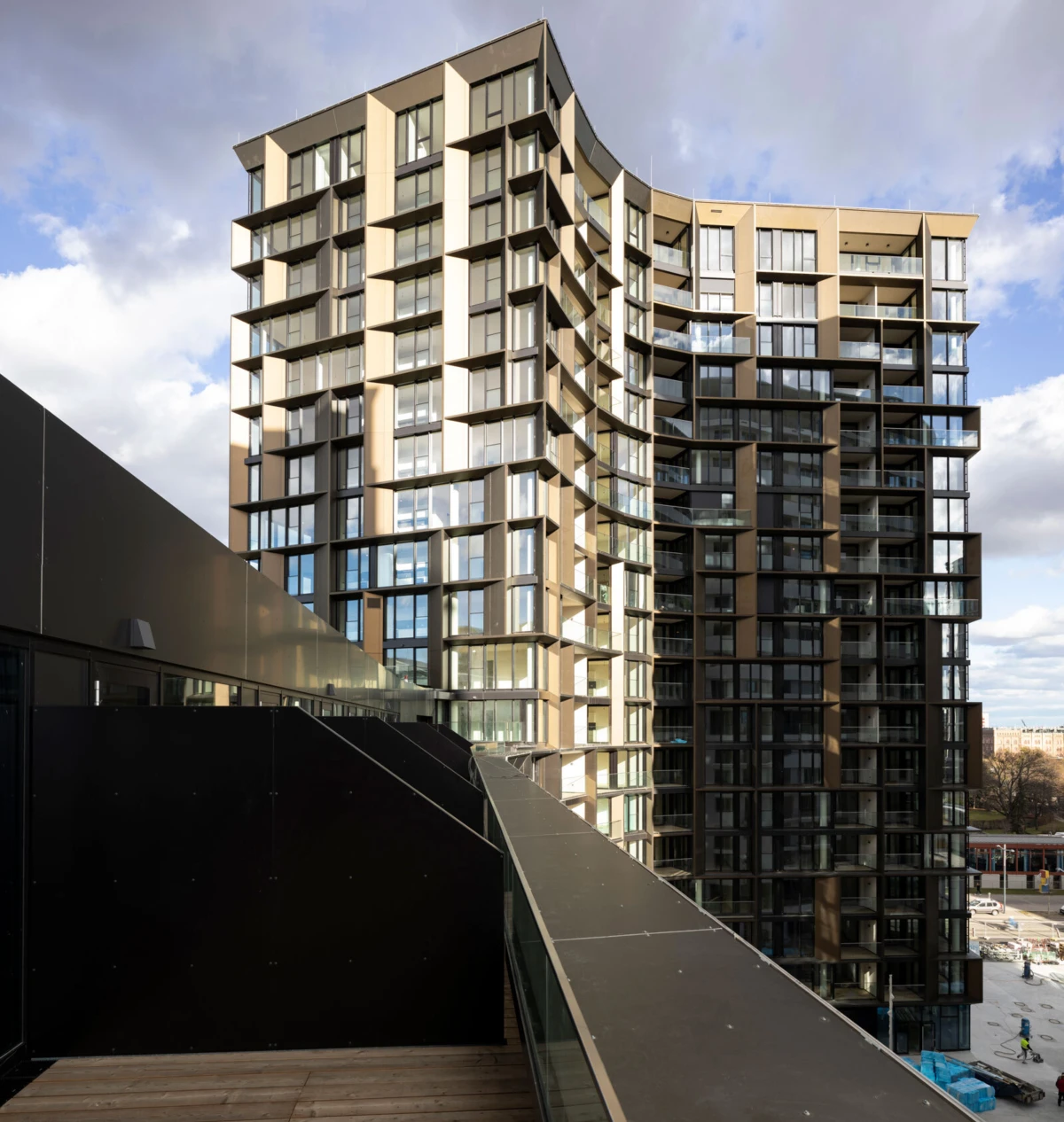 Modern glass-fronted high-rise apartment building with extensive balconies, viewed from a sloped black walkway in an urban setting.