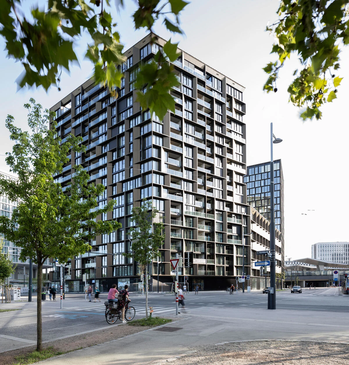 Modern glass-and-concrete high-rise residential tower at a busy city intersection; cyclists and pedestrians on the street beneath trees.