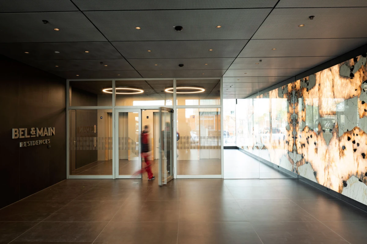 Blurry person walks through glass doors in a modern residential lobby; BEL & MAIN sign on left; leopard-pattern wall art on right.