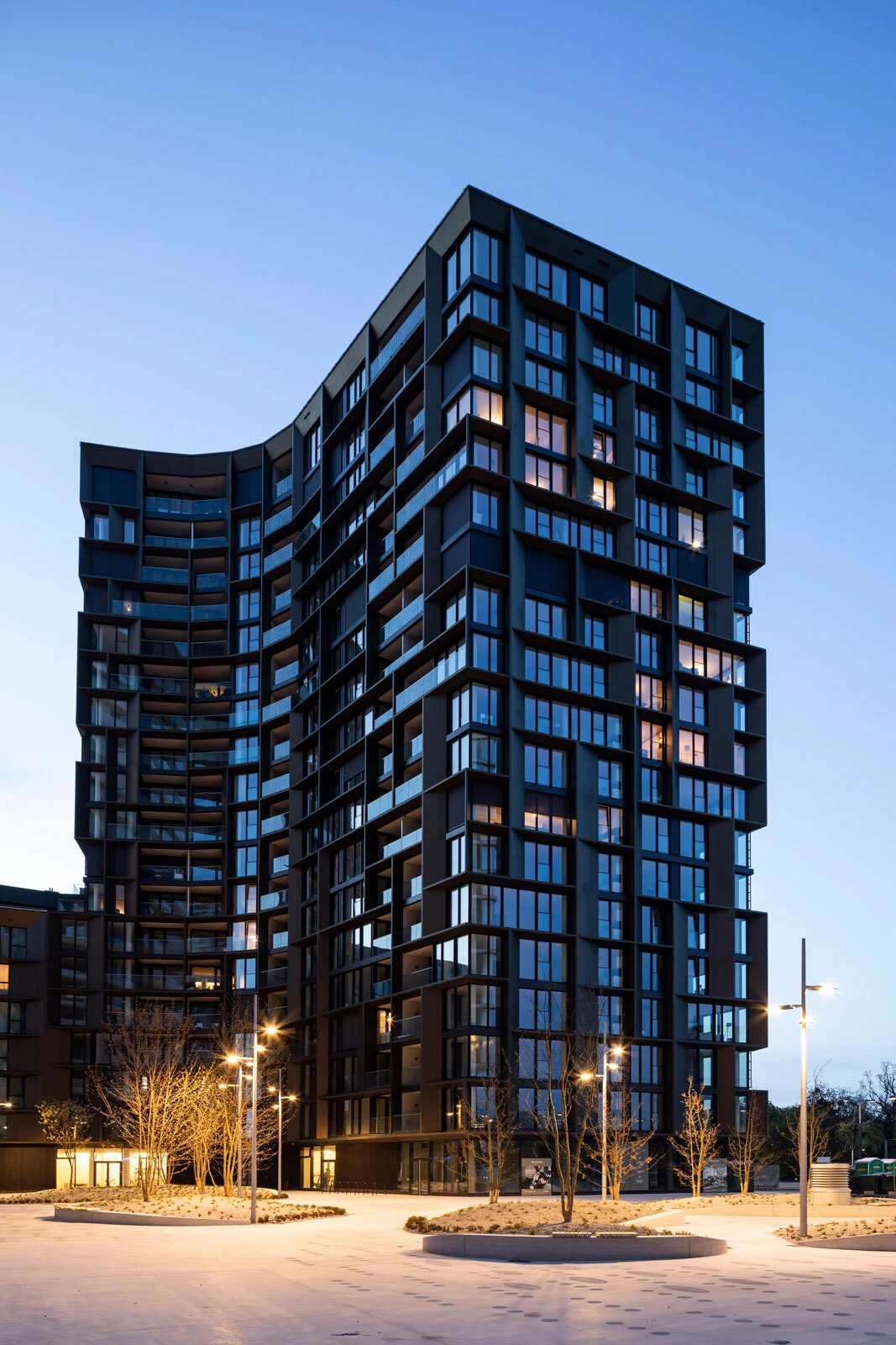 Curved modern high-rise apartment building with lit windows, in a plaza lined with street lamps and bare trees at dusk.