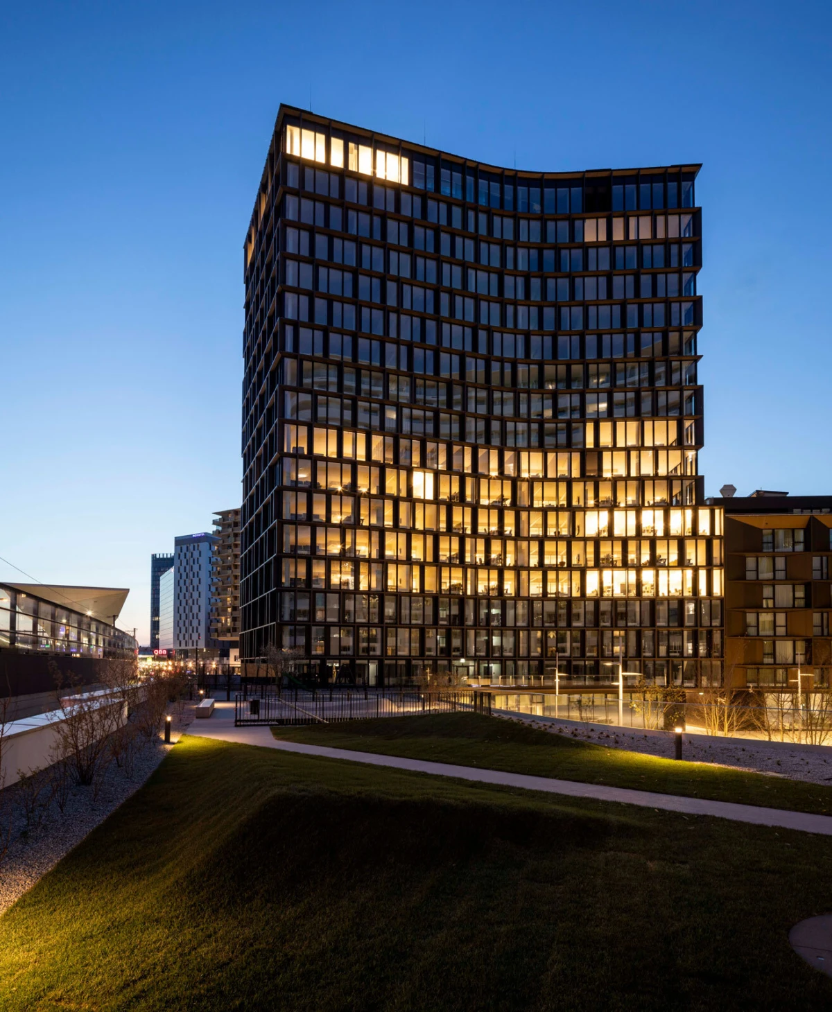 Curved glass-office building with illuminated windows at dusk, set in a campus-like landscape with a walkway and green lawn.