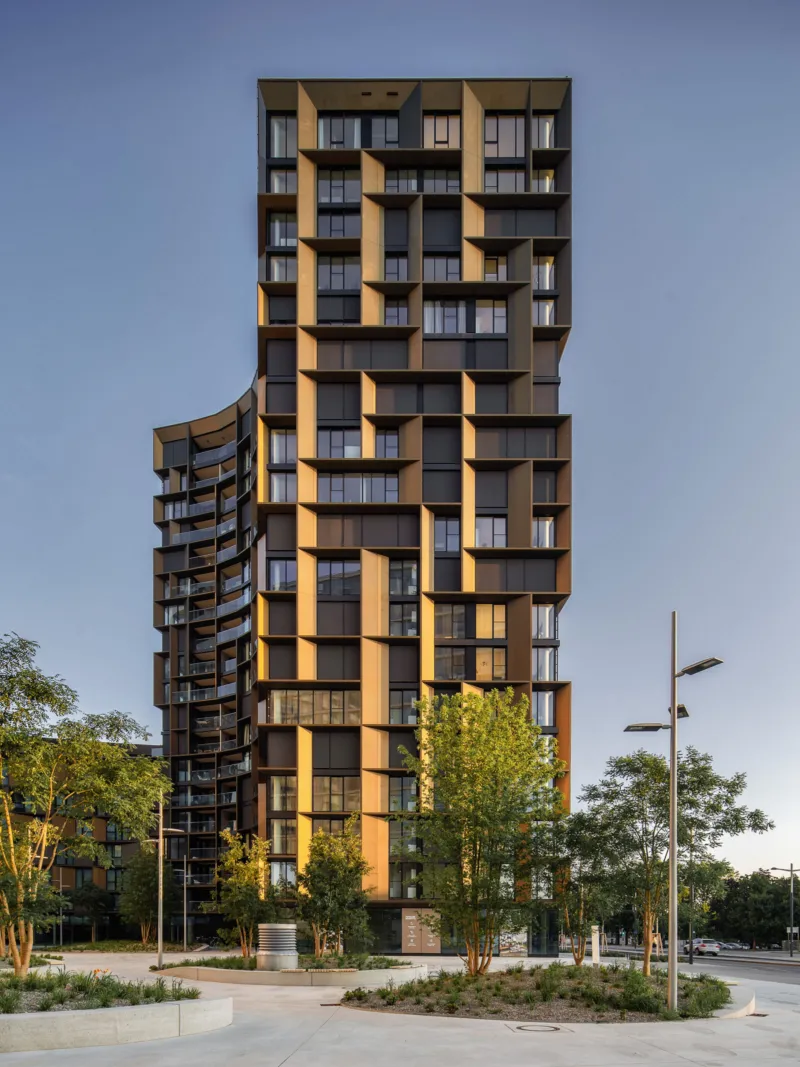 Tall contemporary high-rise apartment tower with stacked box balconies; urban plaza with trees and street lamps.