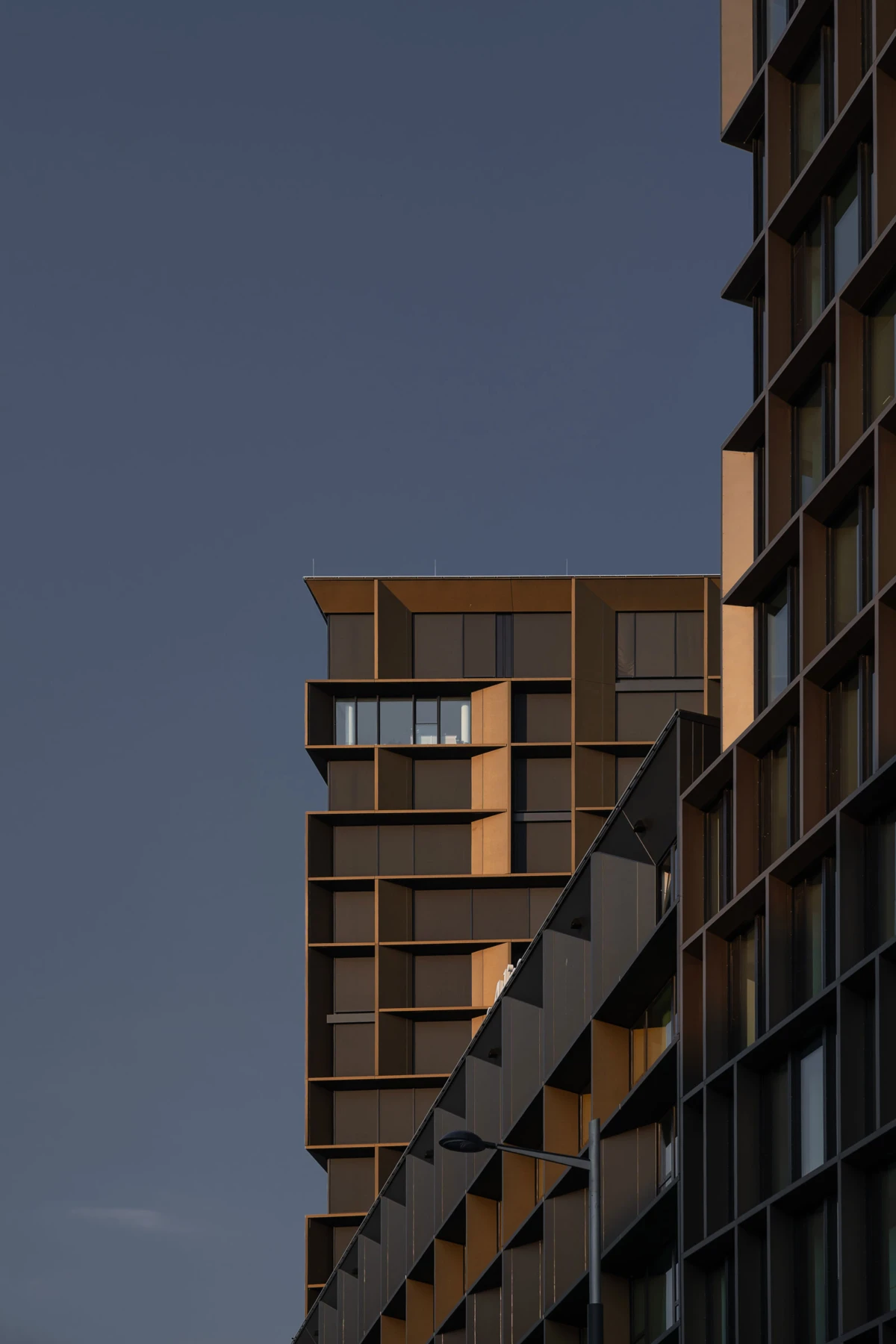Two brown modern apartment towers rise with stepped balconies, connected by a diagonal skybridge, against a clear blue sky.