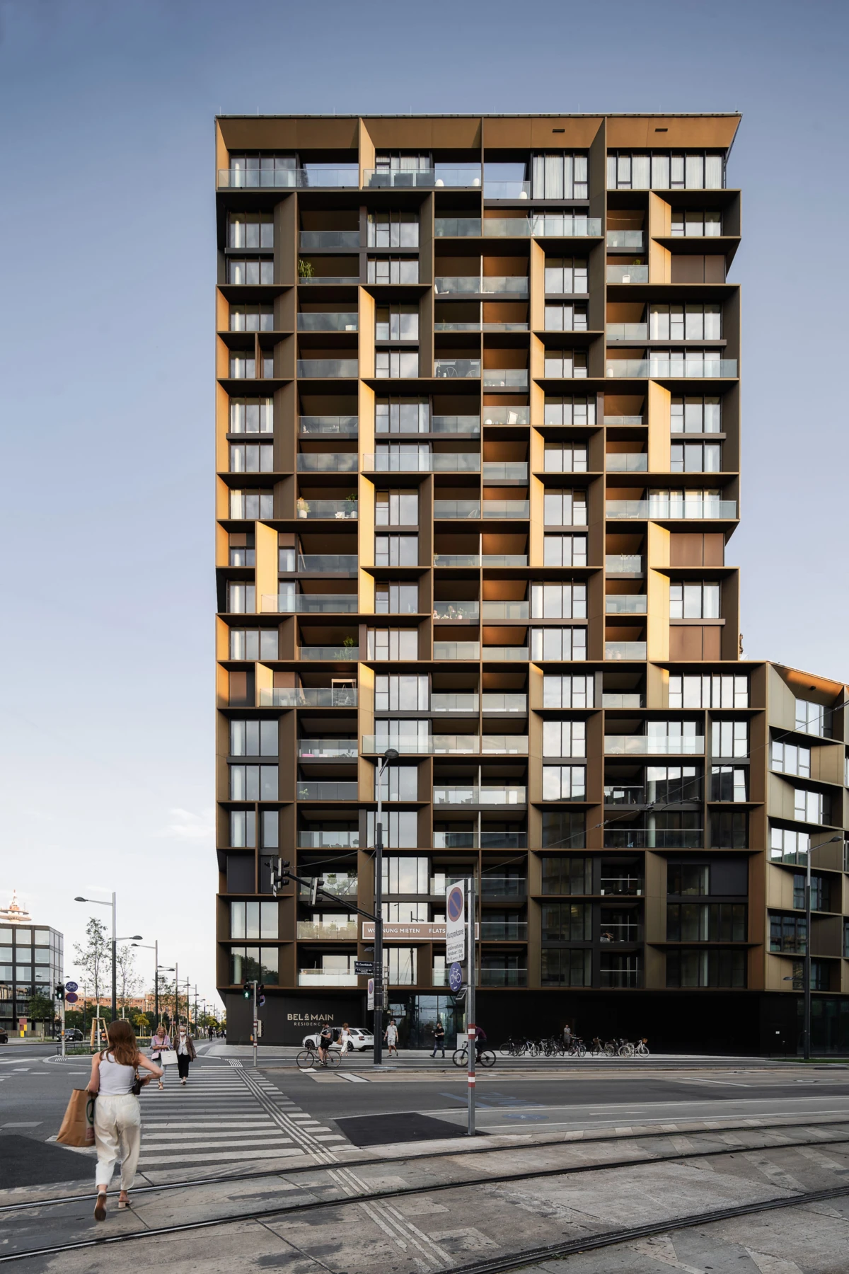 Modern brown-and-black glass apartment building towers over an urban street with tram tracks; pedestrians cross at a zebra crosswalk.