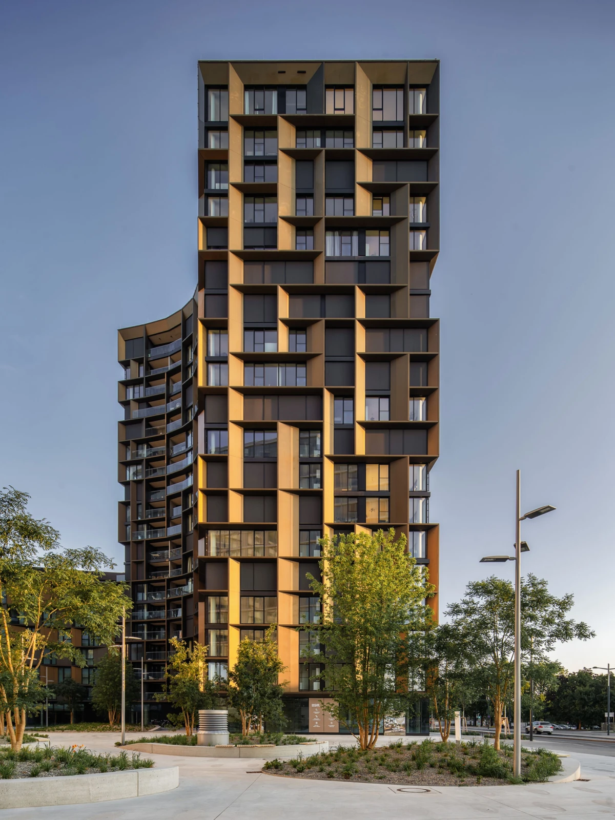 Tall contemporary high-rise apartment tower with stacked box balconies; urban plaza with trees and street lamps.