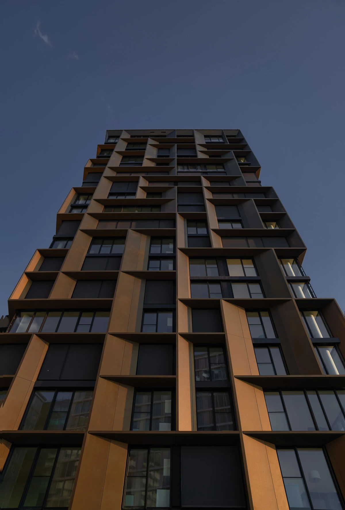 Tall modern high-rise apartment building with orange-brown angular facade and large windows against a blue sky.