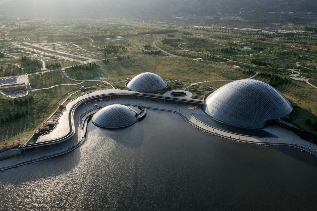 Three glass-domed pavilions connected by a curved walkway over a lake in a large park.