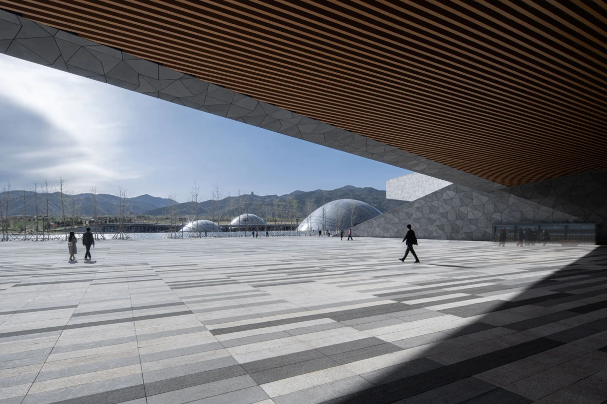 Pedestrians walk across a large tiled plaza beneath a wooden slatted overhang, with geometric walls and glass domes in a mountainous setting.