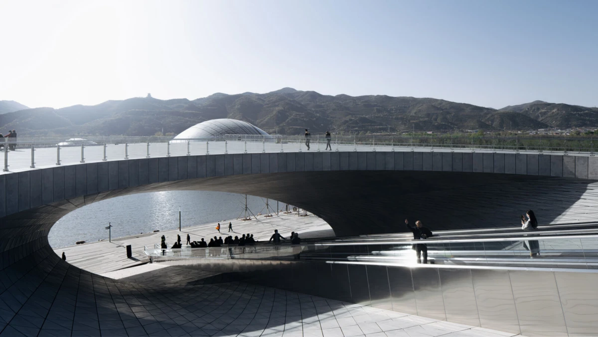 Curved modern pedestrian bridge over water with glass railings; people stroll on the upper deck against a mountainous backdrop.