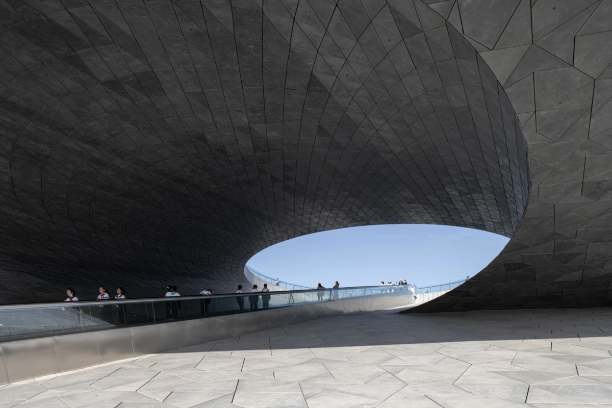 Massive curved dark metal pavilion with a large circular opening; people walk along a glass-railed walkway beneath the overhang, blue sky visible.