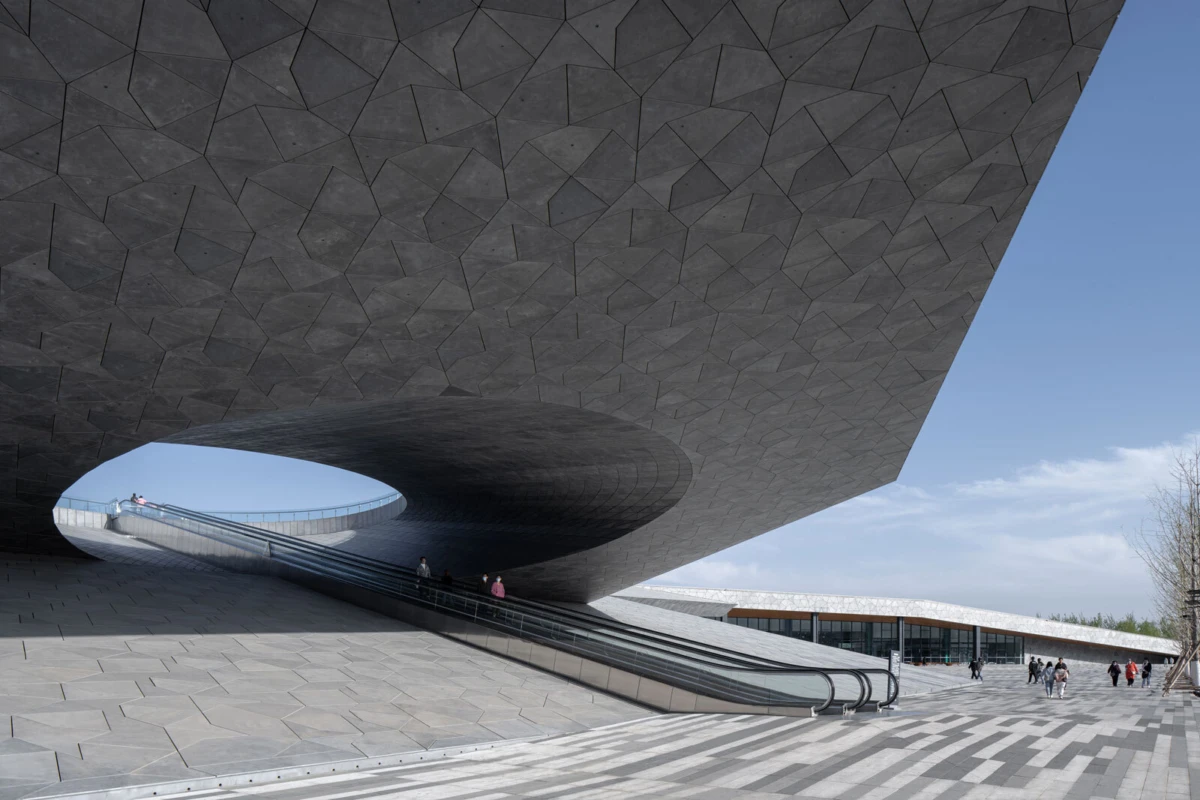Massive concrete pavilion with a circular opening, an escalator, and pedestrians on a tiled plaza beneath a blue sky.