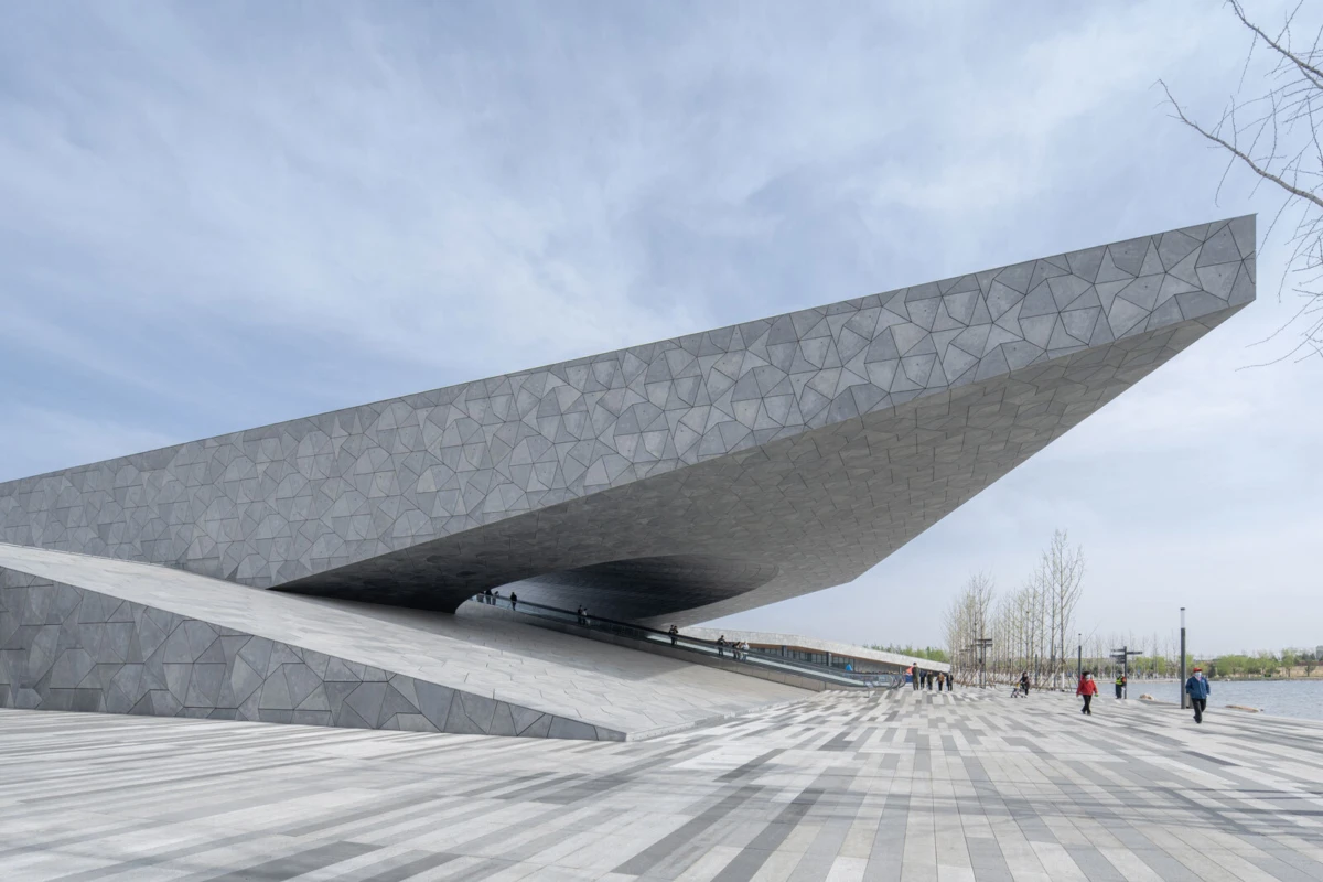 Angular concrete pavilion with a cantilevered roof; pedestrians walk along a tiled promenade beside a lake.