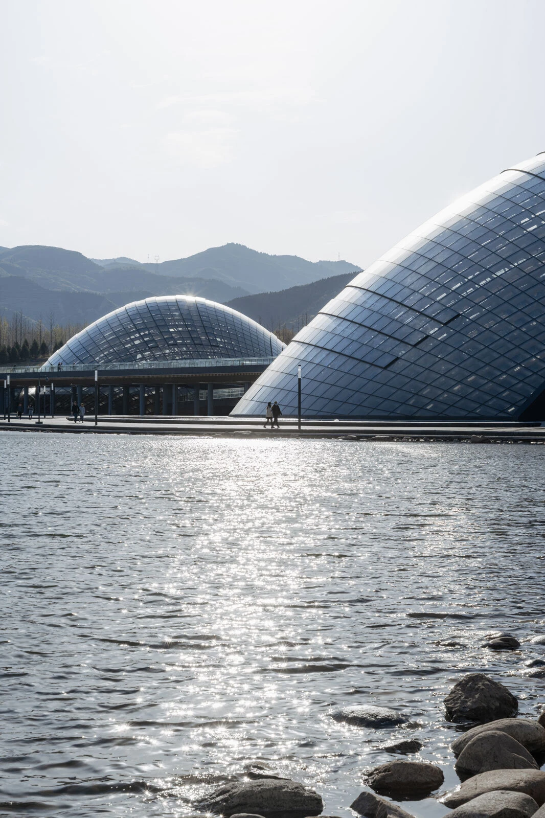 Two large glass-domed structures along a waterfront promenade, with rocky shoreline in the foreground and hills in the background.