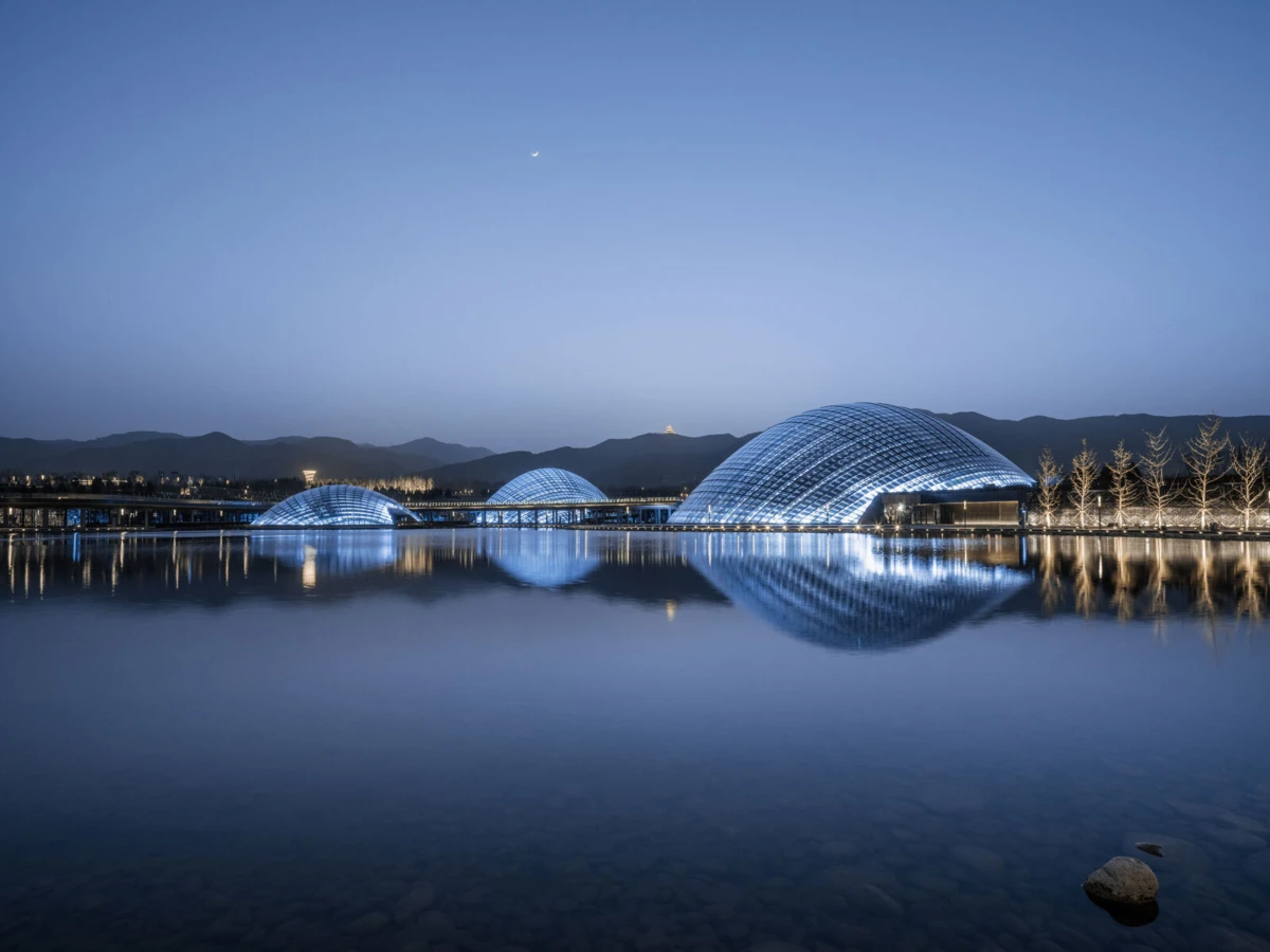Three glass-domed pavilions on a calm lake at dusk, mirrored in the water with a tree-lined promenade and distant mountains.