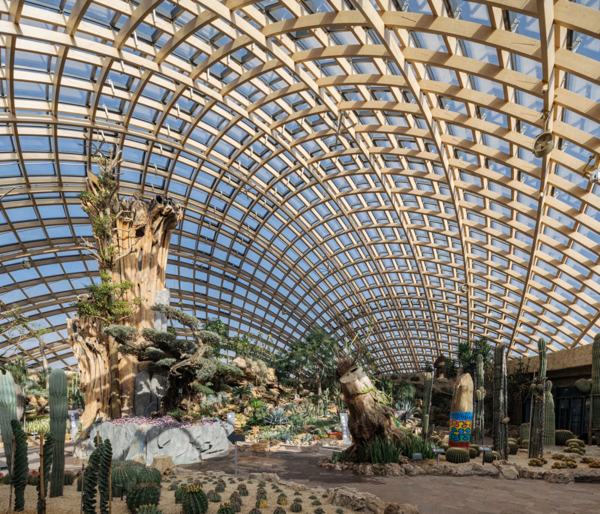 Inside a glass-arched conservatory garden, a tall artificial tree sculpture stands among rocks and cacti.