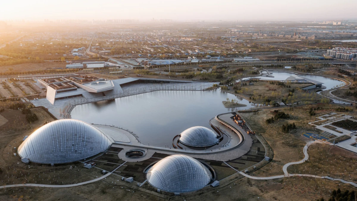 Aerial view of three glass-domed buildings connected by curved walkways around a lake at sunset, with a distant city skyline.