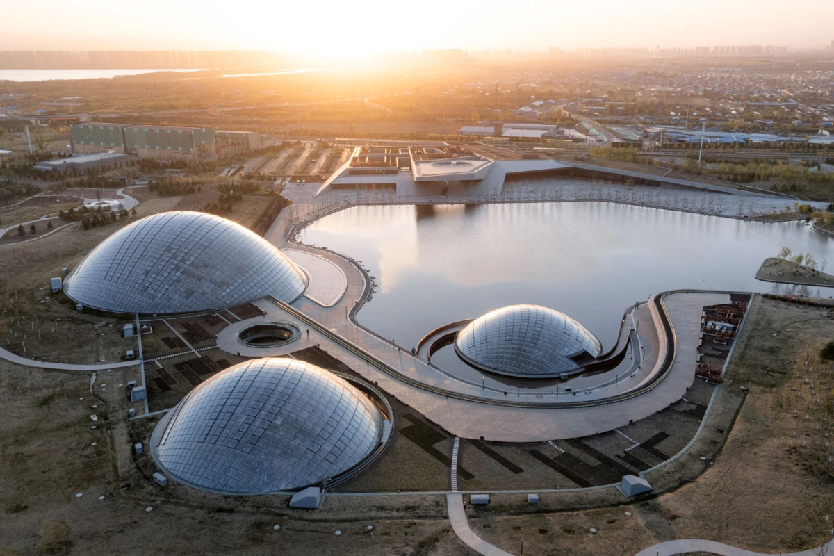 Aerial view of three glass-domed pavilions around a curved walkway by a reflective lake at sunset, with a distant city skyline.
