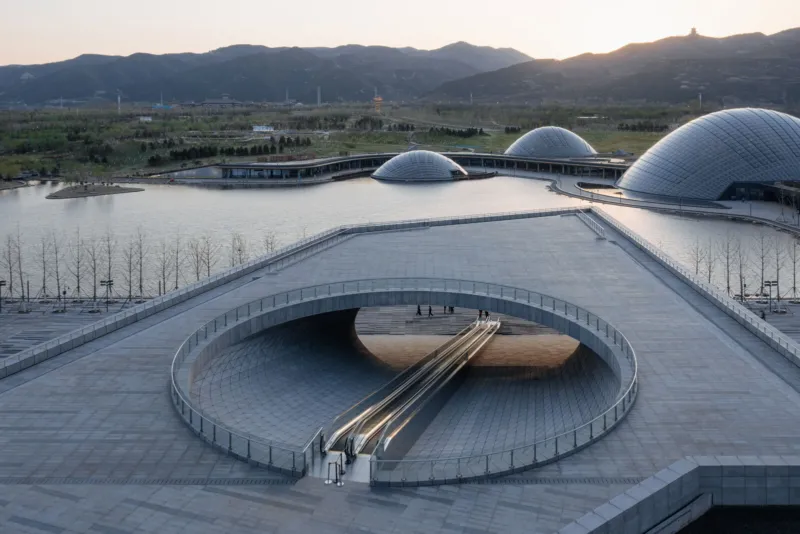 Modern architectural complex with a circular sunken escalator in a ringed opening, gray plaza beside a lake with distant domed pavilions and mountains.