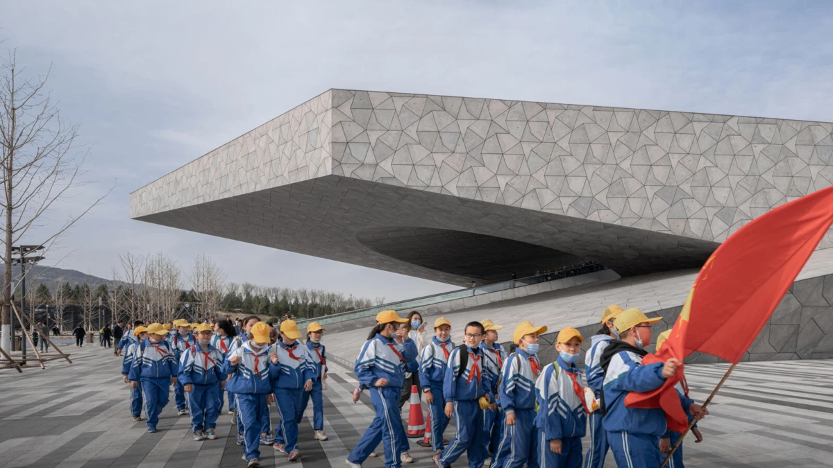 Group of students in blue tracksuits and yellow caps marching with a red flag in a spacious plaza beside a large geometric modern building.
