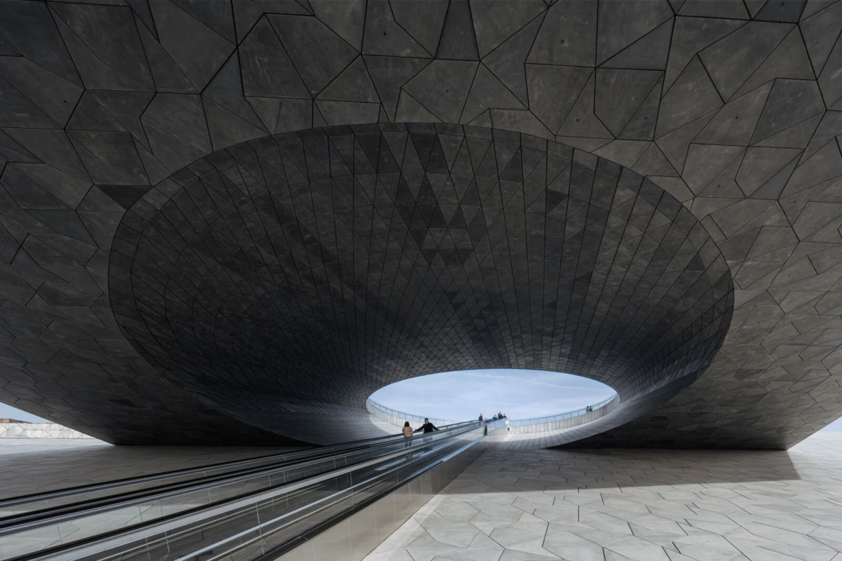 Massive circular opening in a dark geometric ceiling; escalator with people heading toward the bright sky.