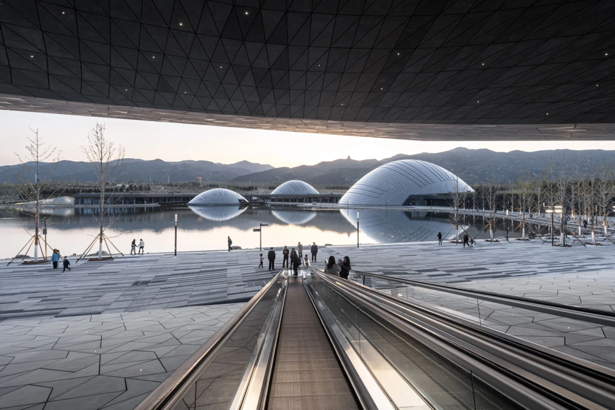 Escalator descending to a plaza by a calm, reflective lake with white domed buildings and distant mountains at dusk.