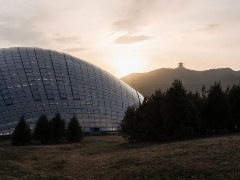 Glass-domed building on a hillside at sunset, with evergreen trees in the foreground and a distant pagoda on the ridge.