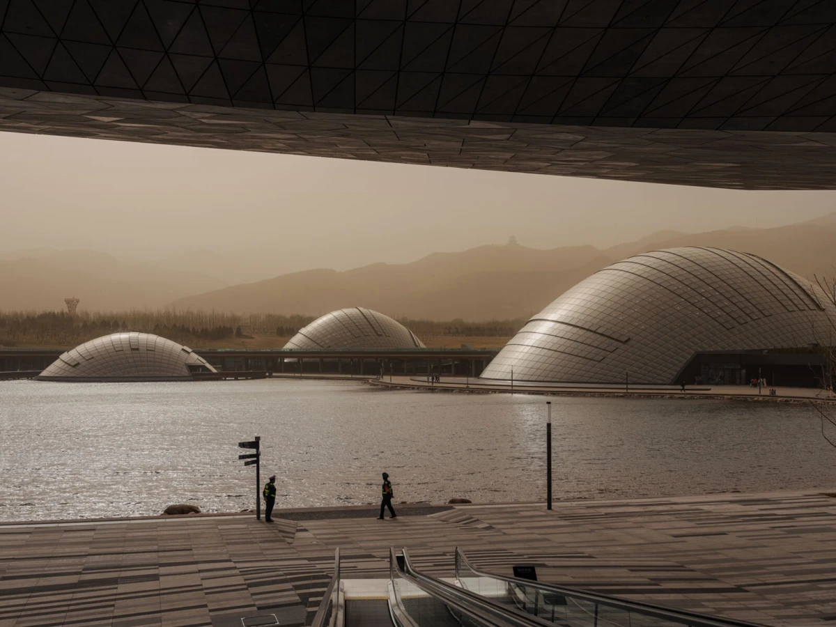 Two workers in reflective vests walk along a lakeside promenade beside futuristic silver geodesic domes on water, under a curved overhanging roof.