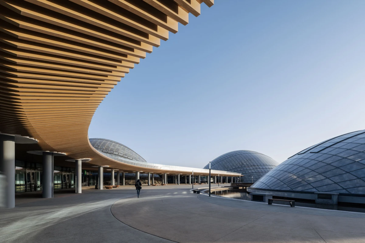 Pedestrian walks along a curved, wooden-slatted promenade beside glass-domed modern architecture on a wide plaza.