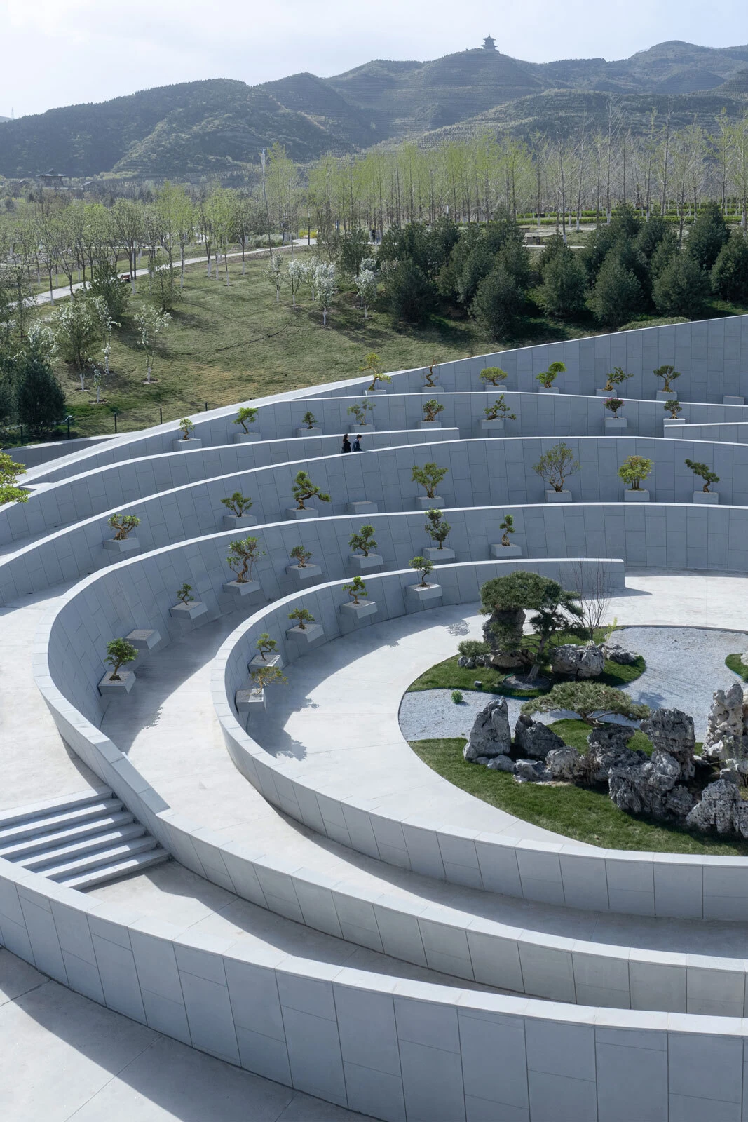 Circular white terraced garden with small potted trees; two people walk the upper level; rocky center with a bonsai tree, hillside park in background.