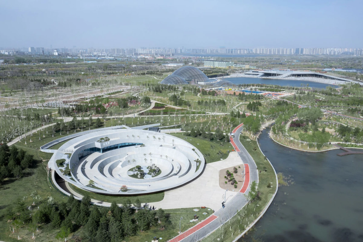 Aerial view of circular white amphitheater in a manicured park along a river, with pedestrians strolling along winding paths.