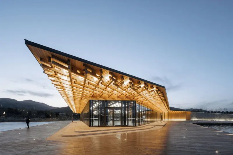 Modern wooden pavilion with an illuminated lattice roof extending over a glass-walled entrance along a waterfront boardwalk at dusk.