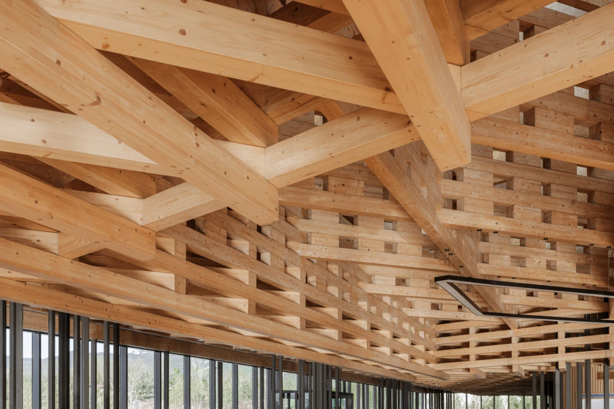 Interior view of an exposed timber-framed ceiling with large crisscrossing wooden beams forming a lattice above windowed walls.