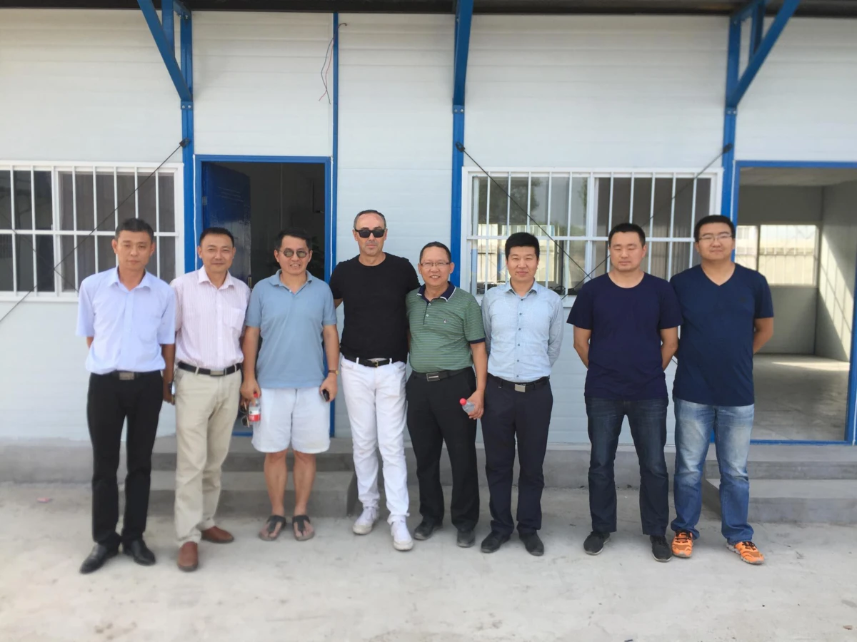Eight men standing side by side, posing for a photo outside a light-gray building with blue columns and barred windows.