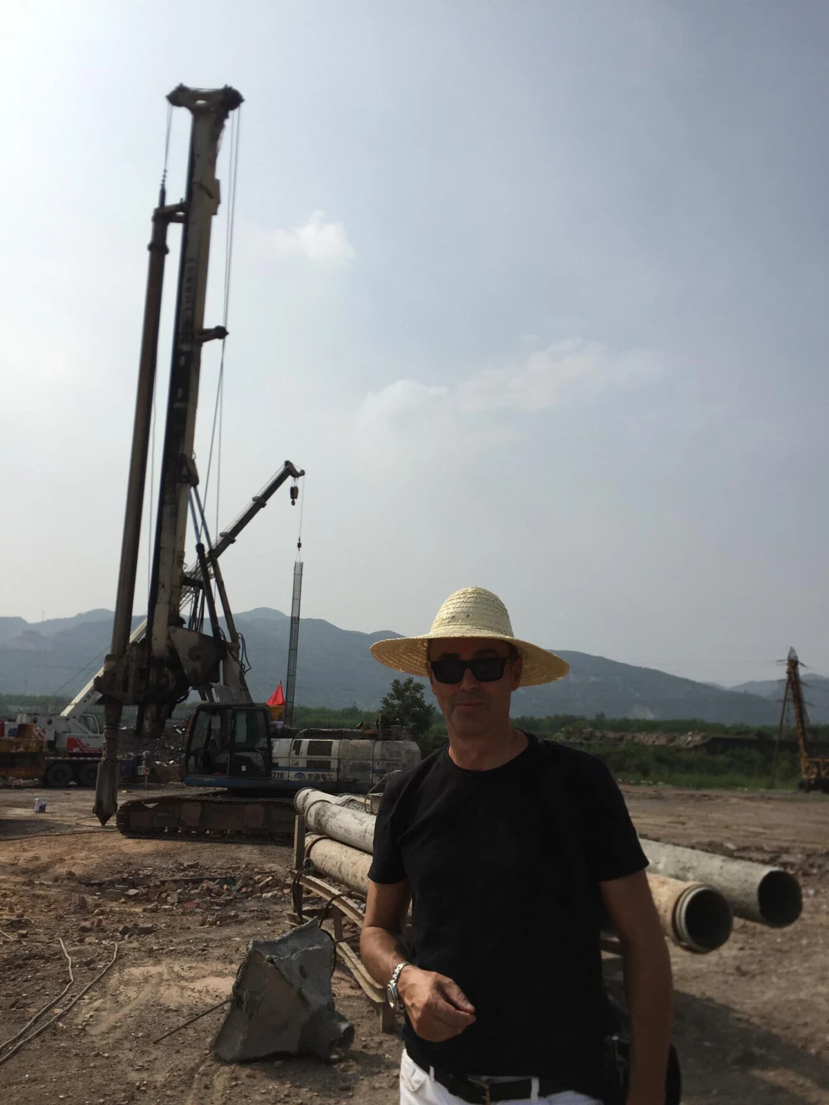 Person wearing straw hat and sunglasses stands at a construction site with a large drilling rig, pipes, and distant mountains.