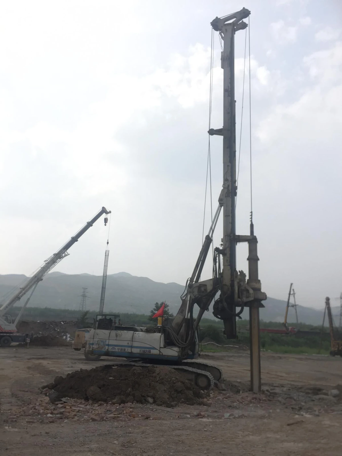 Crawler-mounted drilling rig with a tall derrick drilling into the ground at a construction site; dirt piles and cranes in the background.