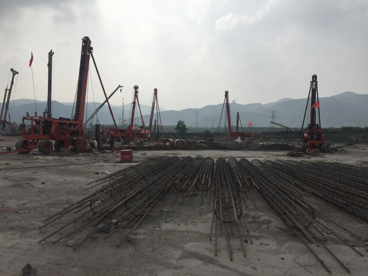 Construction site with several red drilling rigs and workers; bent steel rebar on the ground, mountains in the distance.