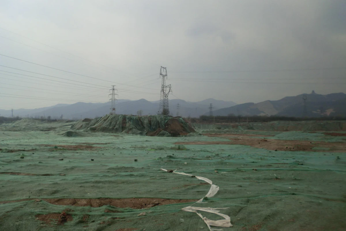 Wide view of an earthworks site with a green tarp-covered mound, power pylons, and distant hills under a cloudy sky.