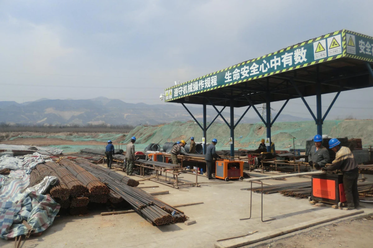 Outdoor construction site with workers in blue helmets handling steel rebar under a safety canopy.