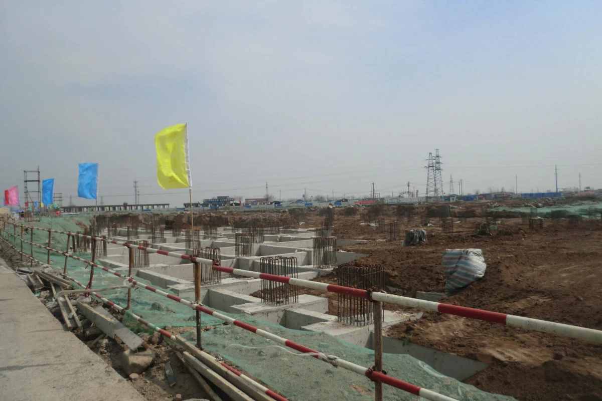 Construction site with concrete footing trenches and steel reinforced bars; colorful flags along a fenced, muddy area under overcast sky.
