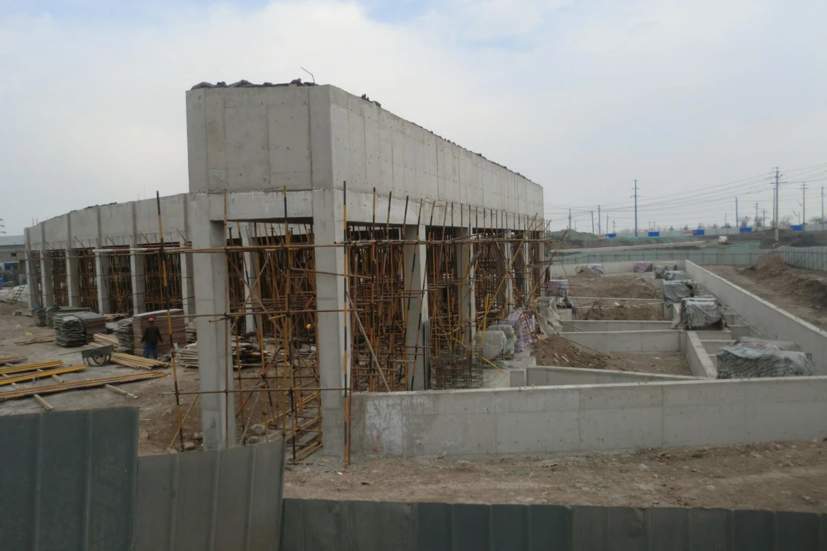 Unfinished concrete building surrounded by scaffolding on a dirt construction site; stacks of lumber and a worker nearby.