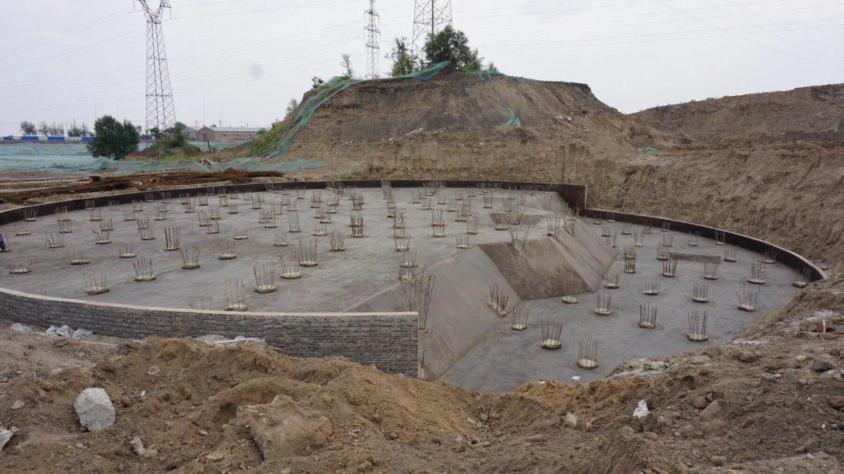 Circular concrete foundation under construction with vertical reinforcement bars; dirt mounds and brick retaining wall surround the site.