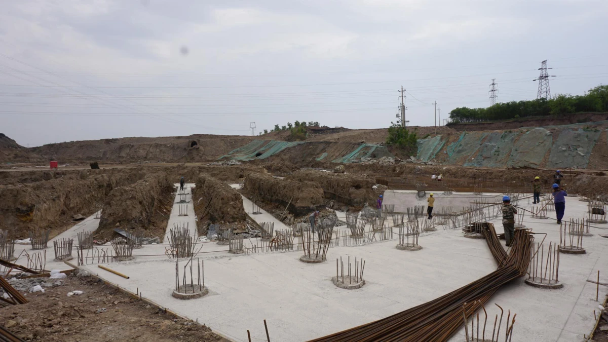Construction site showing circular concrete footings with steel reinforcing bars; workers install foundation amid earth mounds.