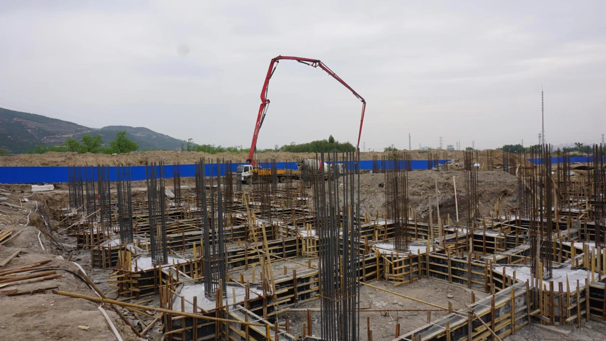 Construction site with vertical rebars and wooden formwork for a reinforced concrete foundation; a concrete pump truck operates in the background.