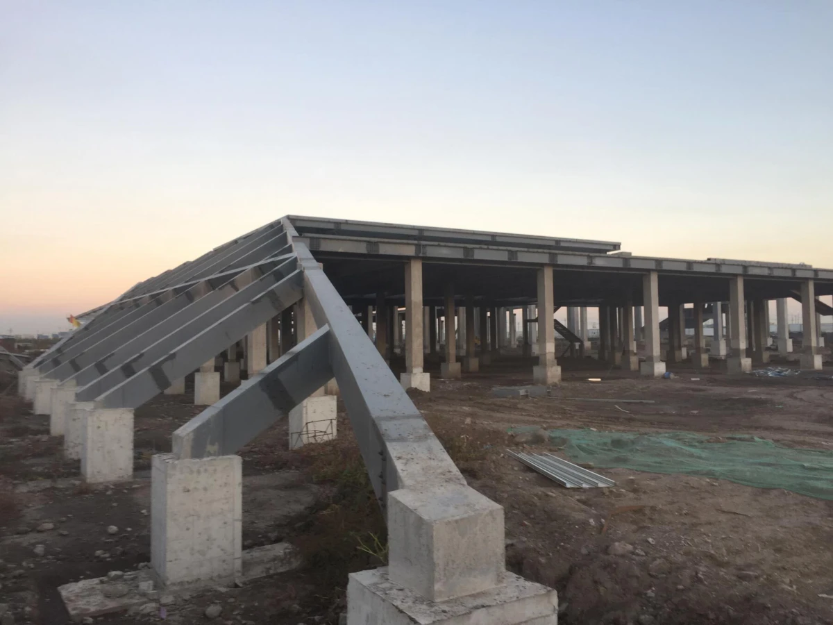 Under-construction concrete-frame building with slanted steel roof trusses on concrete columns; dirt site with scattered metal and netting at sunset.