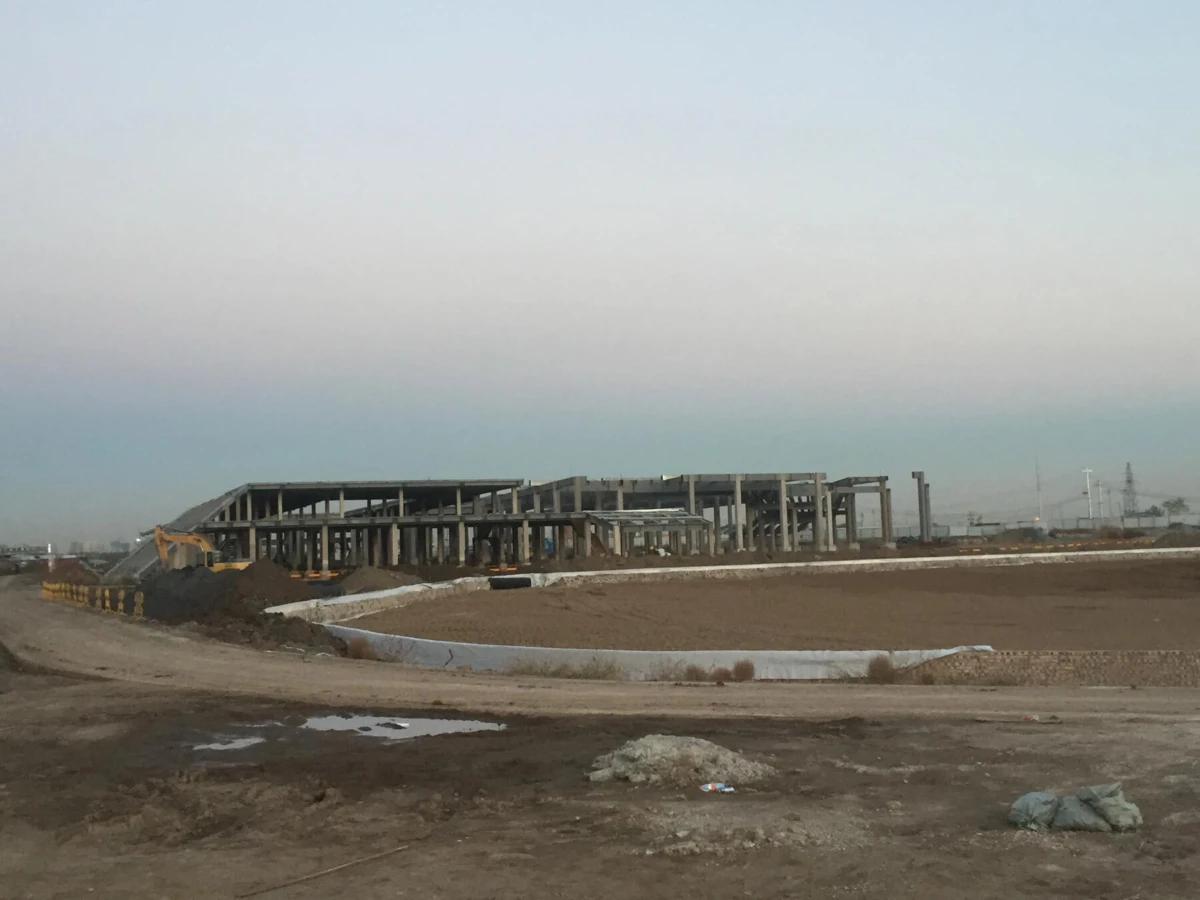 Steel-framed building under construction on a dirt site; excavator on a pile, empty foreground, pale blue sky.