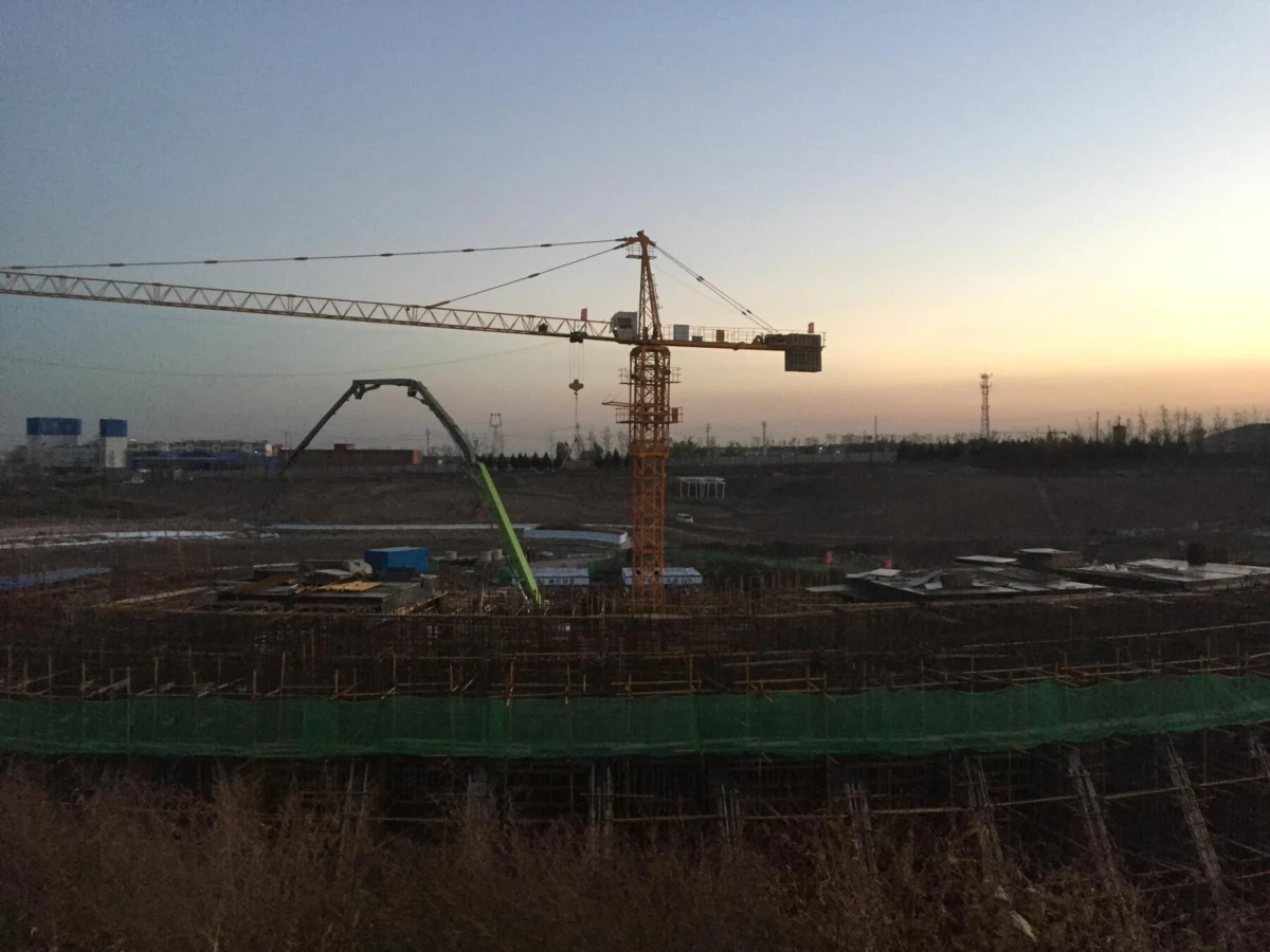 Orange tower crane over a construction site at sunset; foreground shows scaffolding and green safety netting.